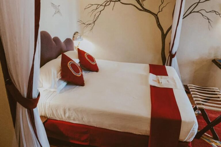 Una cama grande con cojines rojos entre dos cortinas blancas, delante de un árbol pintado en la pared