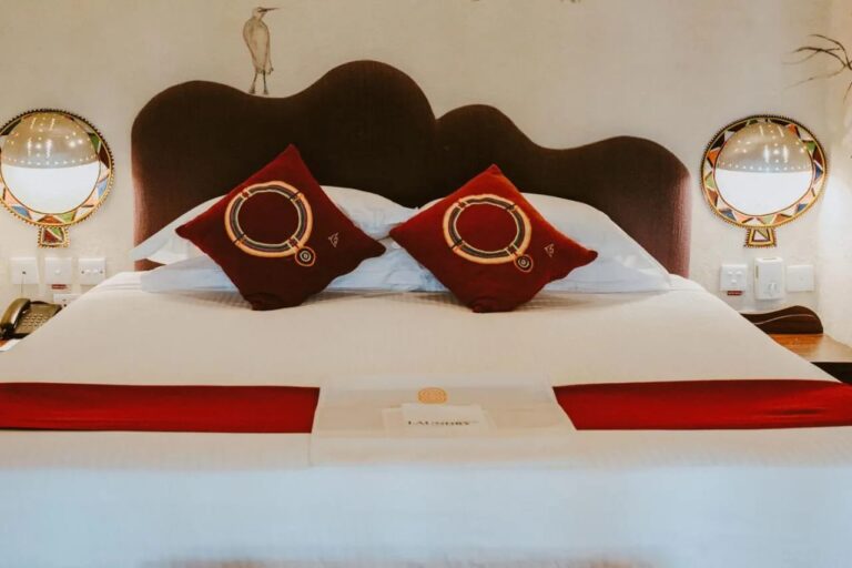 Dos cojines rojos bordados sobre una cama grande, bajo una garza pintada en la pared y entre dos apliques de colores