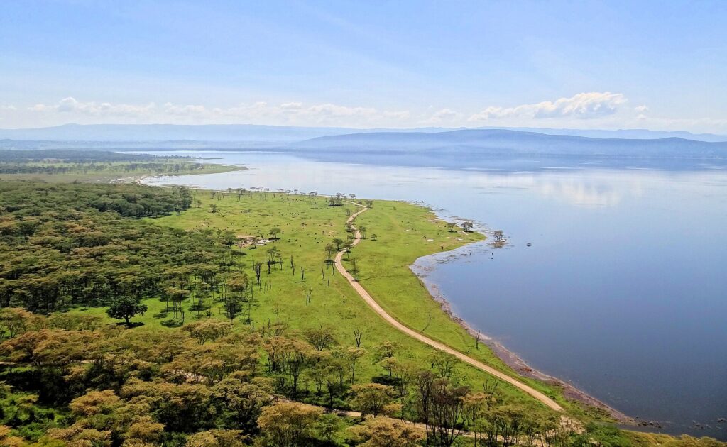 Vista desde lo alto de Baboon Cliff sobre la orilla del lago, con una pista que bordea una pradera verde salpicada de animales, bosques de acacias y el lago hasta las colinas