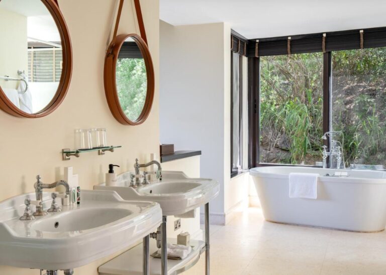 Un baño con dos lavabos de pie, espejos redondos y una bañera delante de un gran ventanal