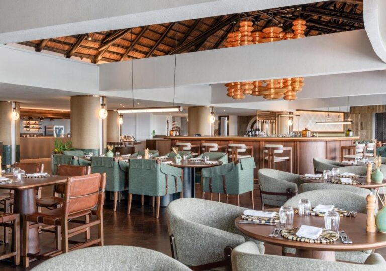 Un restaurante con sillones verdes, delante de una larga barra, bajo lámparas colgantes naranjas