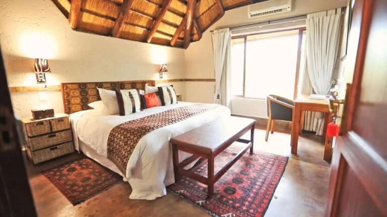 Una habitación bajo una estructura de paja, con una cama grande, un banco de madera y alfombras rojas