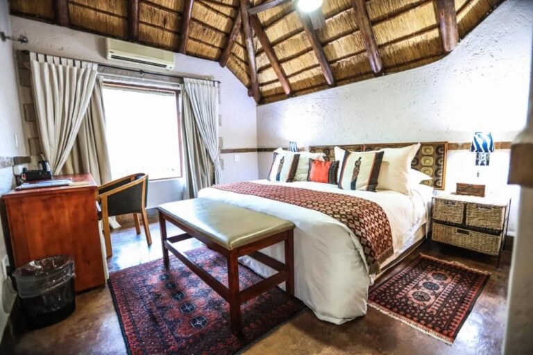 Una habitación bajo el techo de paja, con aire acondicionado, un escritorio de madera y un banco a los pies de la cama