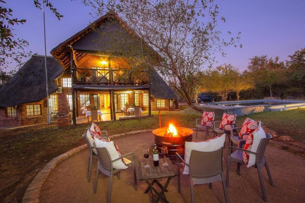 El edificio de ladrillo y paja del Imbube Safari Lodge al anochecer, con un brasero encendido rodeado de sillones, cerca de la piscina