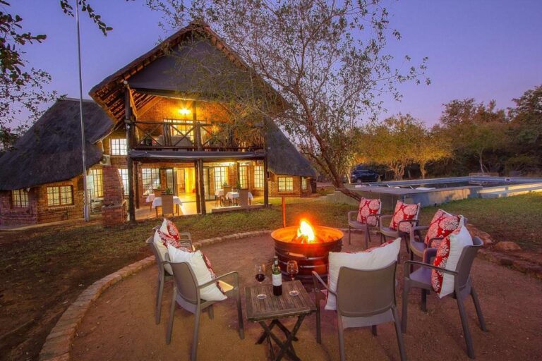El edificio de ladrillo y paja del Imbube Safari Lodge al anochecer, con un brasero encendido rodeado de sillones, cerca de la piscina