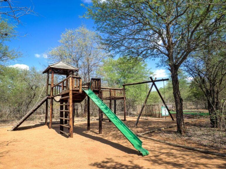 La zona de juegos infantil, con una torre de madera, un tobogán verde y un columpio, bajo los árboles