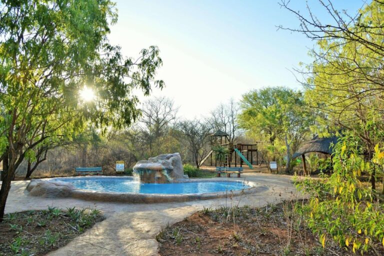 Una piscina con una cascada de rocas, delante de la zona de juegos y una zona cubierta con techo de paja, a contraluz