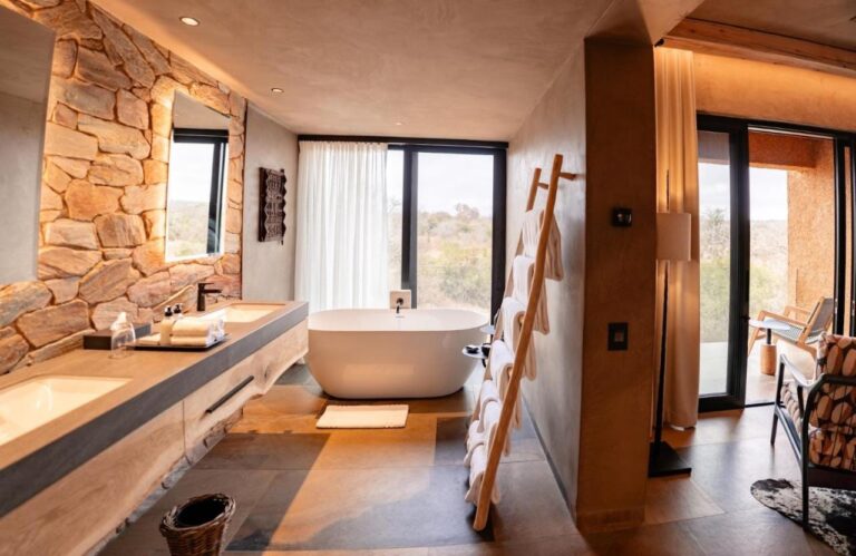 El baño de una suite, con una bañera delante de la ventana y un lavabo doble contra un muro de piedra