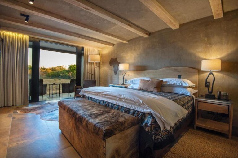 Una suite con cama grande, un banco de piel al pie de la cama y una terraza amueblada frente a la sabana