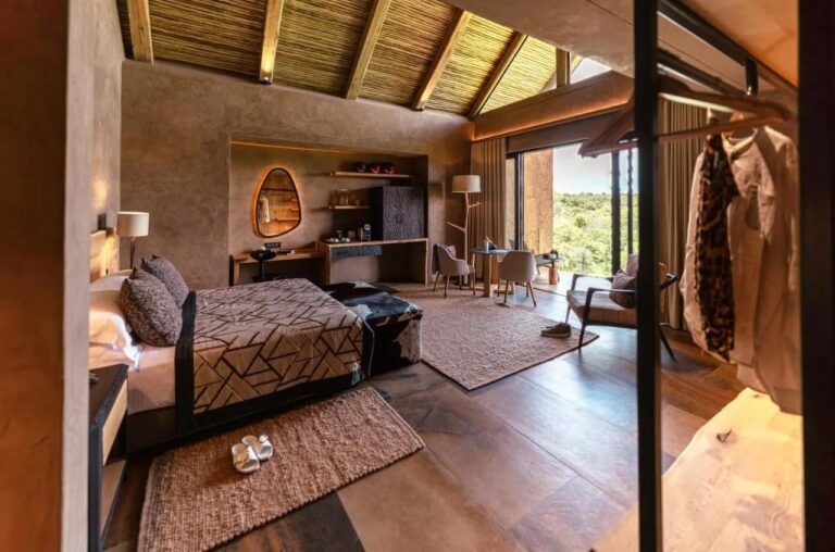Una suite del Kapama River Lodge bajo una estructura de madera y paja, con escritorio y vista a los árboles