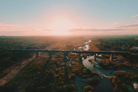 El tren del Kruger Shalati sobre el puente del Selati, visto desde el aire, encima del río y la sabana, con el sol bajo en el horizonte