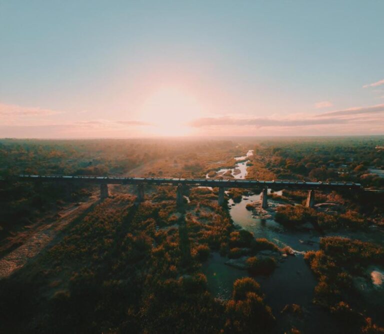 El tren del Kruger Shalati sobre el puente del Selati, visto desde el aire, encima del río y la sabana, con el sol bajo en el horizonte