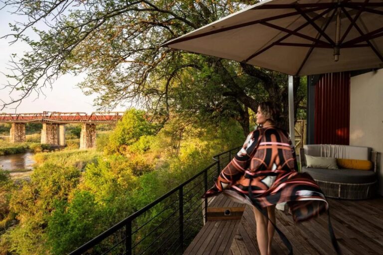 Una mujer en albornoz en una terraza de madera, bajo una sombrilla, frente al puente y al río