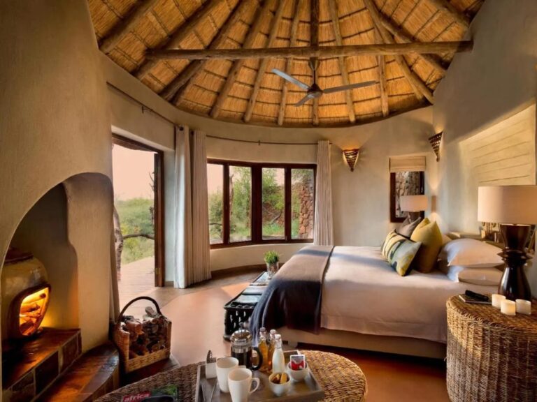 La cama grande de una suite bajo la estructura de paja, entre la chimenea y las ventanas a la sabana