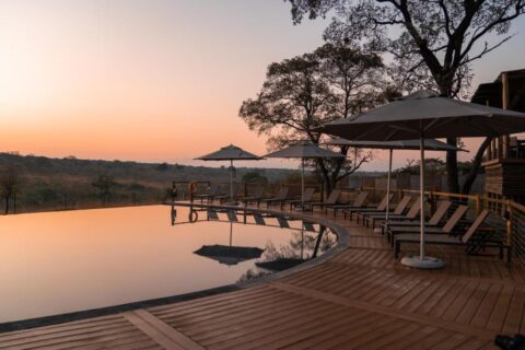 La piscina infinita del Mdluli Safari Lodge bajo un cielo anaranjado, rodeada de tumbonas y sombrillas