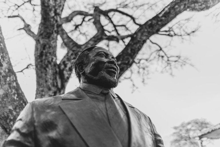 La estatua de bronce de un hombre barbudo y sonriente, de traje, bajo un árbol, en blanco y negro