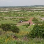 Vista desde Observation Hill de una llanura verde manchada de sal, una pista roja, tres 4x4 estacionados junto a un pequeño edificio ocre y colinas bajas a lo lejos