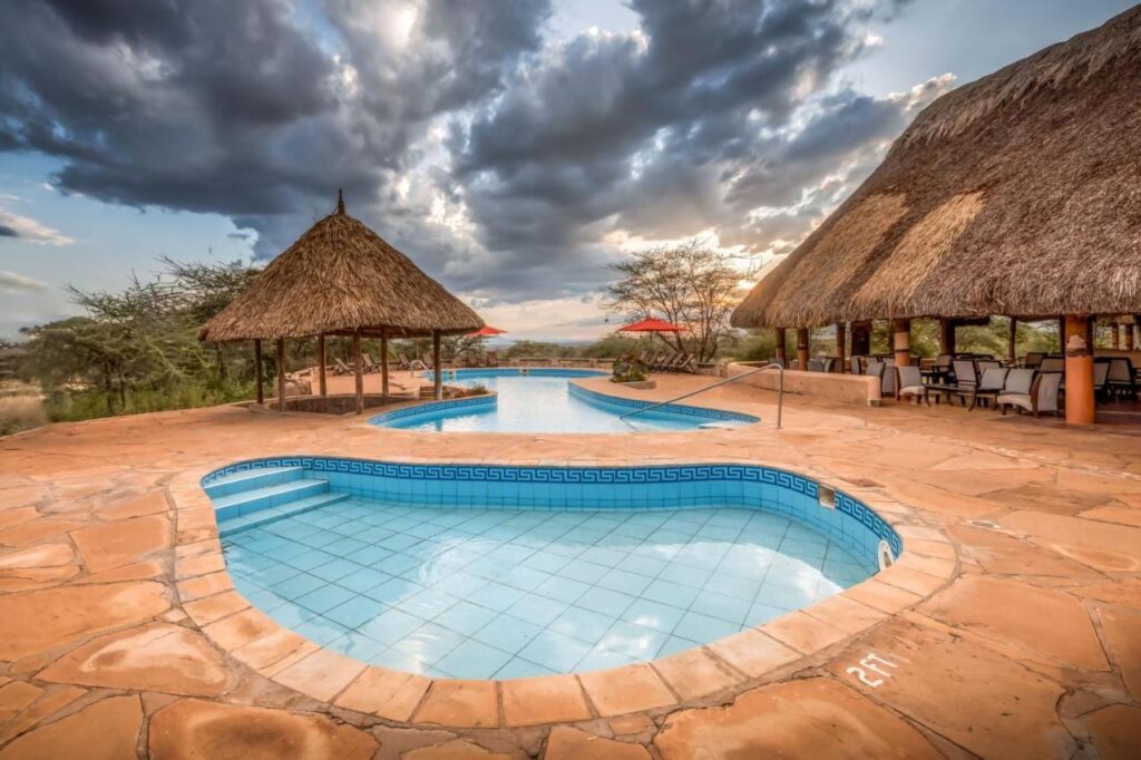 Una pequeña piscina de azulejos en primer plano y la gran piscina del Samburu Sopa Lodge, entre una palapa y una terraza con techo de paja