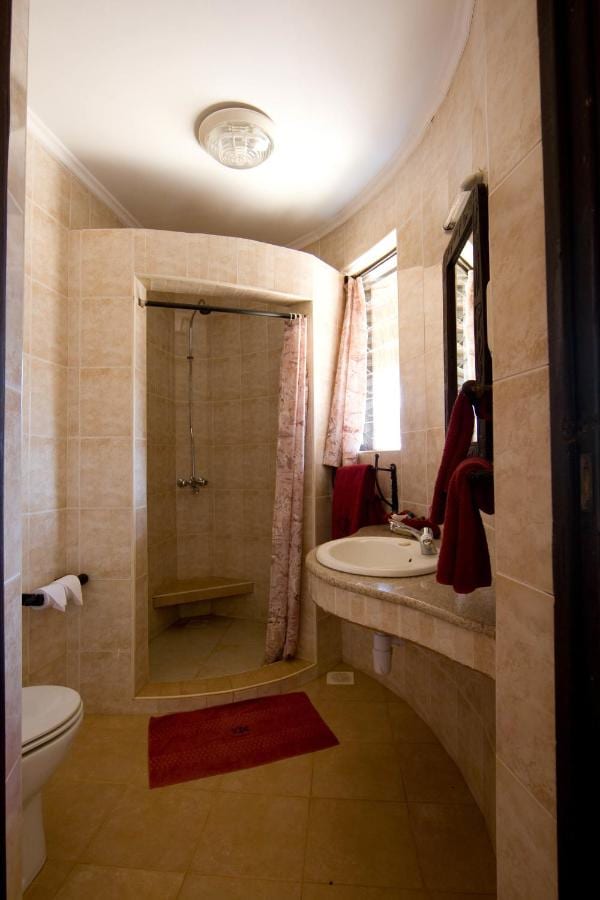 Un baño de azulejos beis, con una ducha con cortina, un lavabo sobre una encimera curva y toallas rojas