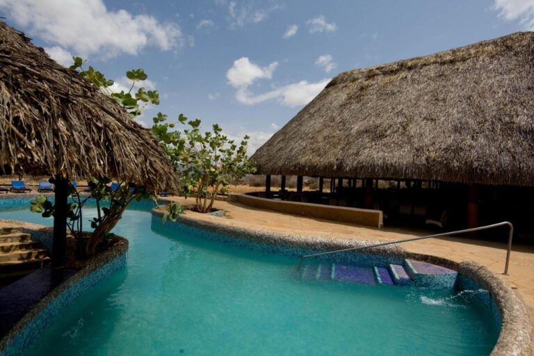 La piscina de mosaico azul, entre una palapa y un gran edificio con techo de paja