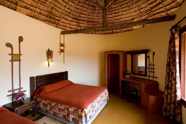 Una habitación con cama grande cubierta de naranja, bajo un techo de paja, con un tocador ocre de formas curvas