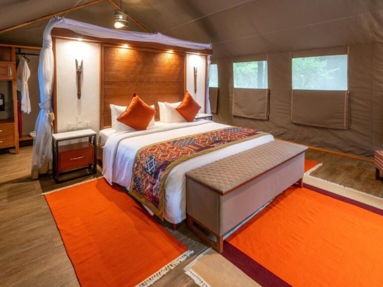 Una tienda con una cama grande bajo mosquitero, cojines naranjas y un banco a los pies de la cama