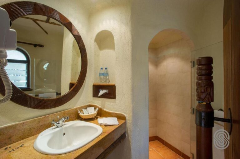 Un baño de paredes enlucidas, con un espejo redondo enmarcado en madera tallada y una ducha bajo un arco