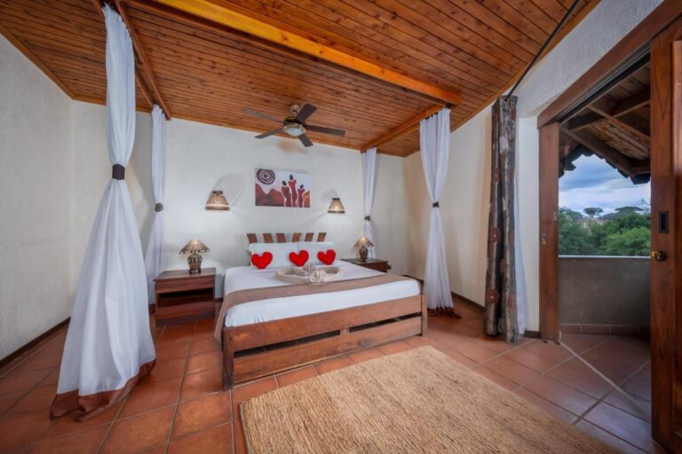 Una habitación con cama grande bajo un techo de madera, con visillos y un balcón