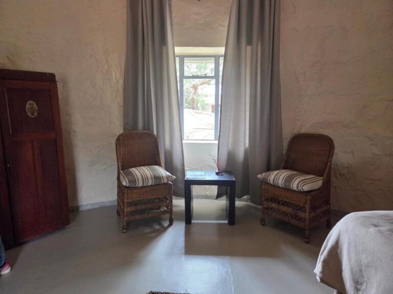 Dos sillones de ratán y una mesita delante de una ventana con cortinas grises, junto a un armario de madera