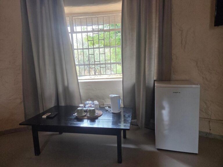 Una mesa baja con tazas y hervidor delante de una ventana con malla, junto a un pequeño refrigerador